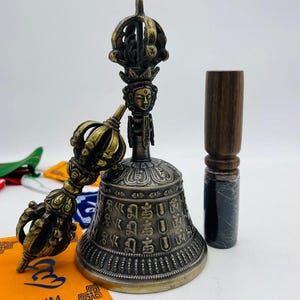 May include: A golden Tibetan prayer bell with intricate carvings and a matching hand-held striker. The bell is decorated with a deity figure and Sanskrit script. A wooden mallet is included.
