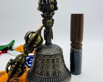 May include: A golden Tibetan prayer bell with intricate carvings and a matching hand-held striker. The bell is decorated with a deity figure and Sanskrit script. A wooden mallet is included.
