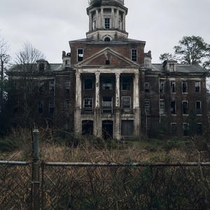 May include: A large, multi-story brick building with a central dome and white columns, showing signs of decay and weathering. The structure features numerous rectangular windows, some broken, and is surrounded by overgrown, dry, brown vegetation.