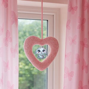 May include: A pink, heart-shaped plush decoration with a small, sparkly bunny charm hanging in the center. The heart is suspended by a pink string, set against a window with pink butterfly curtains.