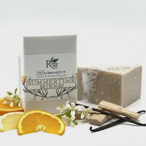 May include: A rectangular bar of beige bath soap with visible bubbles and water droplets, next to a white box labeled Rainwater Botanicals Summertime Bath Soap 5.5oz. The arrangement includes orange slices, white citrus blossoms, and vanilla beans.