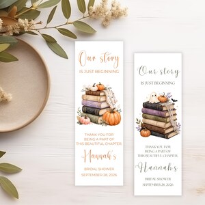 May include: Two rectangular paper bookmarks featuring the text Our story IS JUST BEGINNING, THANK YOU FOR BEING A PART OF THIS BEAUTIFUL CHAPTER, Hannah's BRIDAL SHOWER SEPTEMBER 28, 2026, with watercolor illustrations of stacked books and pumpkins.
