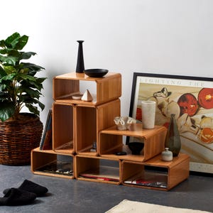 May include: A set of modular, light-brown wooden shelves arranged in a stepped configuration. Various decorative items, including a black vase, small bowls, and a framed print, are displayed on the shelves. A potted plant and a pair of black slippers are also visible.