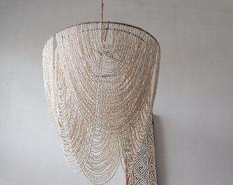 May include: A white shell bead chandelier with a unique cascading design. The chandelier is hanging from a ceiling and is illuminated by a light source. The chandelier is made of many small white beads that are strung together to create a flowing, organic shape.