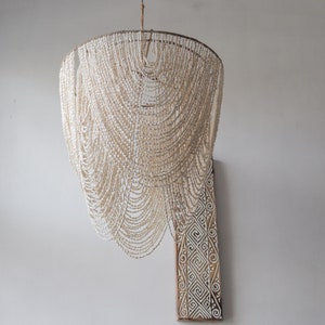 Op de afbeelding: Een witte schelpparel kroonluchter met een uniek cascade-ontwerp. De kroonluchter hangt aan het plafond en wordt verlicht door een lichtbron. De kroonluchter is gemaakt van veel kleine witte parels die aan elkaar geregen zijn om een vloeiende, organische vorm te creëren.