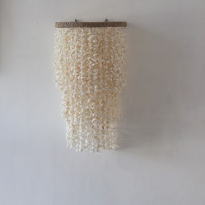 Może przedstawiać: Biały mobil z muszli wiszący na drewnianym pręcie. Mobil wykonany jest z wielu małych białych muszli, które są nawleczone na sznurek w kaskadowym wzorze.