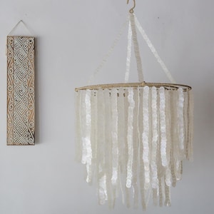 以下が含まれることがあります： 円形のフレームとロープの吊り下げシステムを持つ白い貝殻のシャンデリア。シャンデリアは、カスケード状に垂れ下がった多くの小さな丸い貝殻でできています。