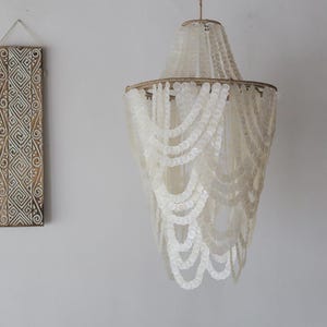 Op de afbeelding: Een witte schelpenkroonluchter met een rond frame en meerdere strengen schelpen die eraf hangen. De kroonluchter is gemaakt van natuurlijke materialen en heeft een rustieke, bohemian stijl.