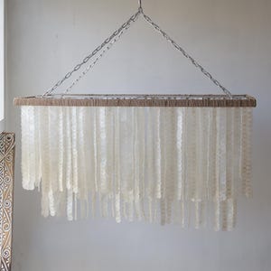 May include: A rectangular chandelier made of natural materials. The chandelier features a woven jute frame and cascading strands of white, round, shell-like beads.