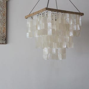 May include: A white shell chandelier with a square wooden frame and a rope trim. The chandelier is made of many small, rectangular shells that hang in tiers.