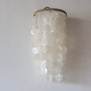 May include: A cylindrical hanging light fitting featuring multiple tiers of translucent, circular capiz shell discs suspended from a circular frame wrapped in natural tan jute twine. The white shell discs overlap to create a layered, textured appearance.