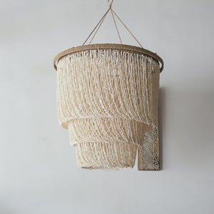 Puede incluir: Una lámpara de araña escalonada con una estética natural y bohemia. La lámpara presenta un marco circular envuelto en un material marrón claro, con hebras de cuentas blanquecinas en cascada. Cuelga de un cordón de fibra natural.