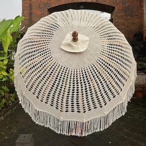 Può includere: Un parasole color crema a trama aperta con manico in legno e finiture a frange. La capote del parasole presenta un design circolare con un terminale centrale in legno e una frangia decorativa sul bordo. La trama aperta lascia filtrare la luce.