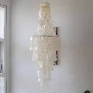 Può includere: Un lampadario di conchiglie bianche con più livelli di conchiglie rotonde appese a una struttura metallica. Il lampadario è montato su una parete.