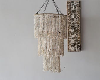 Op de afbeelding: Een witte kralen kroonluchter met drie lagen, hangend aan een ketting tegen een witte muur.