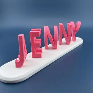 May include: A pink 3D-printed nameplate that spells out the name "JENNY". The letters are in a bold, sans-serif font and are mounted on a white, oval-shaped base. The background is a solid, dark blue.
