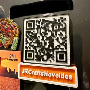 May include: A square, 3D-printed magnet featuring a black and white QR code pattern. Below the QR code is a rectangular orange base with white, raised lettering that reads JRCraftsNovelties, all set against a speckled white background.