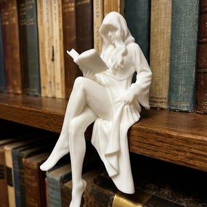 May include: A white 3D printed figurine of a hooded figure with long hair sitting on an edge whilst reading an open book. The figure wears a long, flowing robe with wide sleeves and is positioned with legs crossed, showcasing a smooth, matte finish.