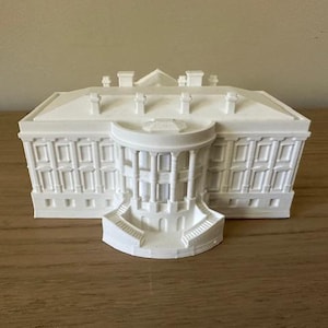 May include: A white miniature replica of the White House. The model features detailed architectural elements, including columns, windows, and a central portico with stairs. The building is set on a wooden surface.