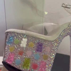 Könnte beinhalten: Transparenter Plateau-High-Heel-Schuh mit Strasssteinen auf Basis und Absatz. Die Basis ist mit bunten Gummibärchen- und sternförmigen Verzierungen verziert. Der Schuh hat einen transparenten Knöchelriemen.