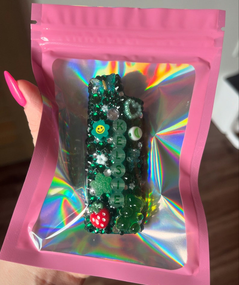 Green and silver rhinestoned lighter cover saying “Kelsie” in green letters. Features a butterfly charm, a pearl heart, a smiling flower, a sour gummy star, a strawberry, and a gummy bear.