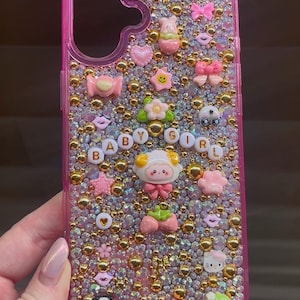 May include: A pink phone case decorated with numerous iridescent crystals, gold beads, and various miniature charms including bows, hearts, stars, strawberries, and a central sheep figure. The text BABY GIRL is spelled out in white letter beads.