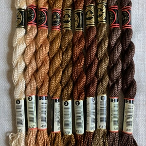 Op de afbeelding: Een verzameling DMC parel katoenen borduurgaren in verschillende tinten bruin, beige en goud. Elke streng is voorzien van een nummer en merkinformatie. Het garen is strak opgerold en klaar voor handwerkprojecten.