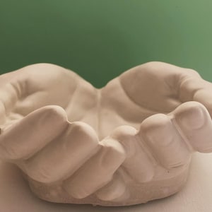 May include: A cream-colored, open-hand sculpture, designed to cradle objects. The hands are sculpted with detailed fingers and a textured surface, creating a bowl-like shape. The background is a solid green.