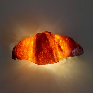 May include: A glowing, golden-brown croissant-shaped light fixture. The light emanates from within, highlighting the textured surface and shape of the pastry. The fixture is suspended against a neutral background.