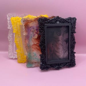 May include: A collection of ornate picture frames in various colors, including clear, yellow, and black. The frames have detailed, decorative edges and are arranged in a row against a pink background. The frames are empty.