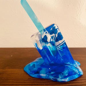 May include: A translucent blue popsicle sculpture featuring a light blue stick and a melting base. The popsicle body displays a white and blue floral pattern embedded within the clear resin, creating a realistic appearance of a frozen treat.