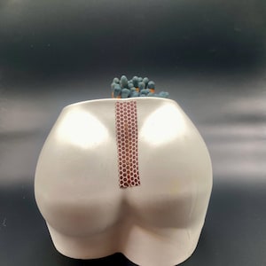 May include: A white ceramic match holder shaped like a backside, with a strip of match striker paper down the center. The match holder is filled with blue-tipped matches.
