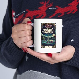 Peut inclure: Une tasse en céramique blanche avec une illustration colorée d'une fleur blanche sur un fond bleu foncé. Le texte "Juillet" est imprimé sous l'image.