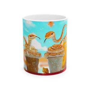 May include: A white ceramic mug featuring a colourful illustration of two snakes wrapped around cups of coffee. The snakes are orange and brown with black eyes and smiling faces. The background is a light blue sky with white clouds and falling leaves.