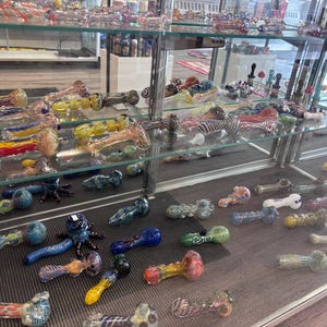 Puede incluir: Una vitrina llena de pipas de vidrio de colores. Las pipas vienen en varias formas y tamaños, con una gama de colores que incluyen azul, amarillo, rojo y verde. Algunas pipas tienen diseños y patrones intrincados.