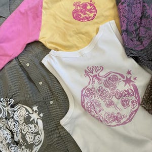 May include: A collection of garments including a white ribbed tank top with a pink pomegranate graphic, a yellow long-sleeve shirt with pink sleeves and a pink pomegranate print, and two button-down shirts with pomegranate graphics.