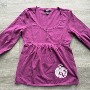 May include: A long-sleeved magenta top with a scoop neckline and a three-button placket. The garment features a gathered empire waist and a white line-art graphic of a pomegranate with a small star on the lower right side. The label reads Style & Co.