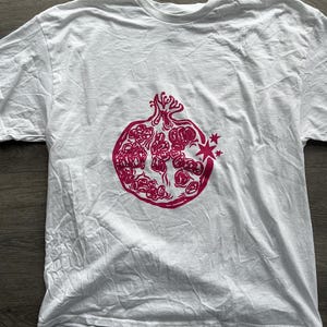 May include: A white short-sleeved t-shirt featuring a centered graphic print of a pomegranate in a vibrant magenta color. The pomegranate illustration shows the fruit split open to reveal its seeds, with a small starburst detail on the right side.