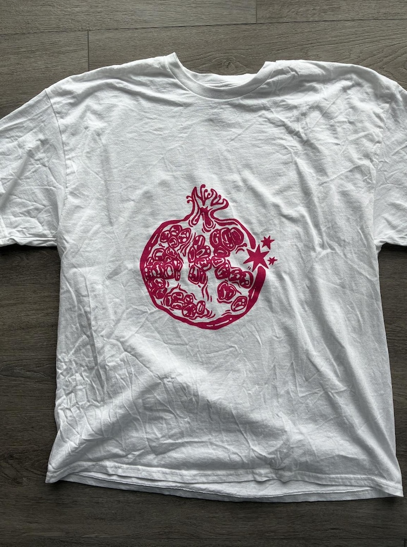 May include: A white short-sleeved t-shirt featuring a centered graphic print of a pomegranate in a vibrant magenta color. The pomegranate illustration shows the fruit split open to reveal its seeds, with a small starburst detail on the right side.