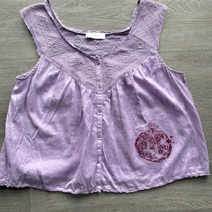 May include: A sleeveless lavender button-down top featuring a square neckline with white geometric embroidery. The garment has a relaxed fit and a dark purple graphic print of a pomegranate on the lower right side with a raw, frayed hemline.