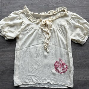 May include: A cream-colored short-sleeve blouse featuring a ruffled collar with a central tie closure. The garment has puff sleeves and a red graphic print of a pomegranate fruit located on the lower right side of the front panel.