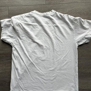 May include: A plain white short-sleeved t-shirt with a crew neckline. The garment is made of a solid white fabric and features standard stitching along the sleeves and hemline. The shirt is displayed flat, showing the back side of the garment.
