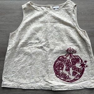 May include: A sleeveless beige top with a textured fabric finish and a round neckline. The garment features a large, dark maroon graphic print of a pomegranate on the lower right side, accompanied by a small star icon near the fruit's edge.