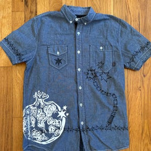 May include: A blue short-sleeve button-down shirt featuring a white pomegranate graphic, a black flail illustration, and black barbed wire patterns on the sleeves and hem. The shirt includes two chest pockets and white buttons.