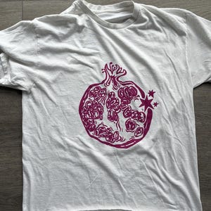 May include: A white short-sleeved t-shirt featuring a magenta graphic print of a pomegranate on the front. The pomegranate illustration includes stylized seeds and two small four-pointed stars on the right side of the fruit.