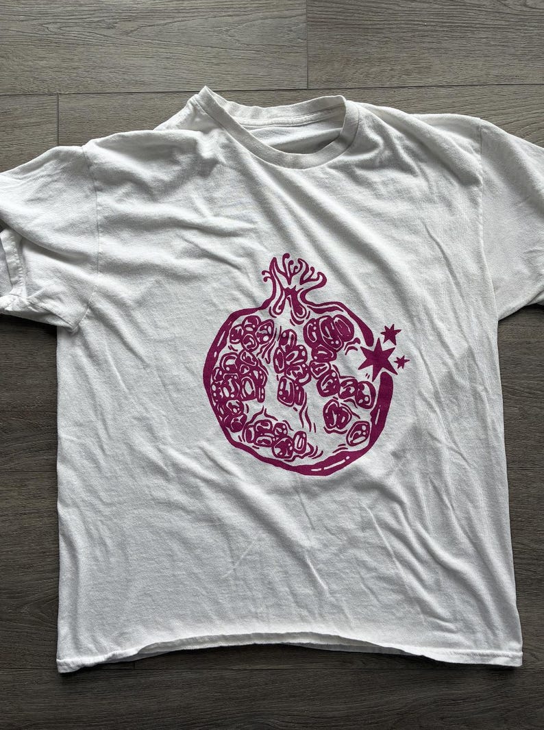 May include: A white short-sleeved t-shirt featuring a magenta graphic print of a pomegranate on the front. The pomegranate illustration includes stylized seeds and two small four-pointed stars on the right side of the fruit.