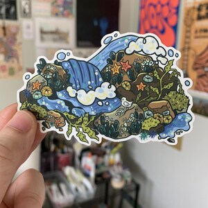 May include: A colorful sticker featuring a waterfall scene with a variety of marine life. The design includes a waterfall, starfish, seaweed, and a small bird. The sticker has a white border and is held by a hand.