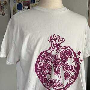 May include: A white short-sleeved t-shirt featuring a large, dark magenta graphic print of a halved pomegranate on the front. The pomegranate illustration includes detailed seeds and two small four-pointed stars positioned to the right side.