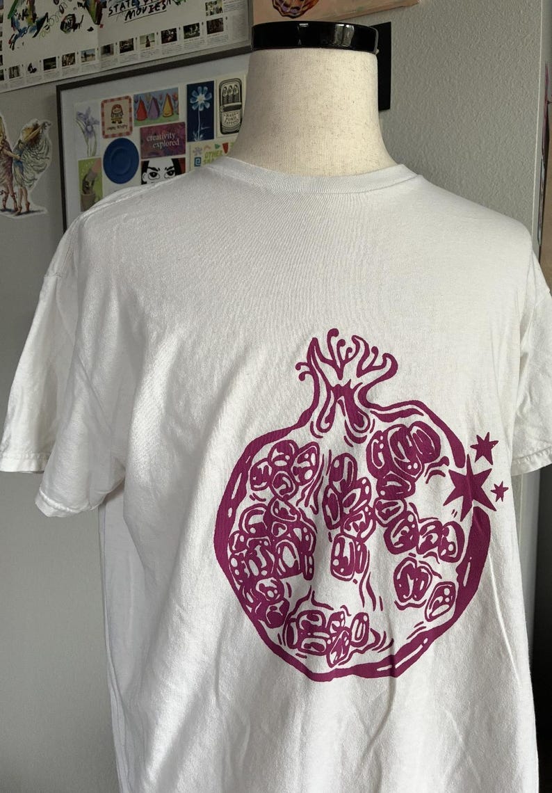 May include: A white short-sleeved t-shirt featuring a large, dark magenta graphic print of a halved pomegranate on the front. The pomegranate illustration includes detailed seeds and two small four-pointed stars positioned to the right side.