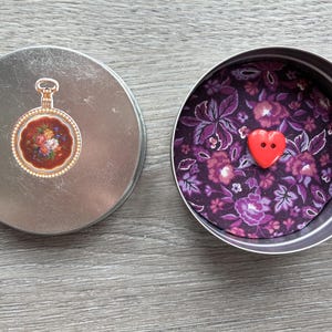Può includere: Due barattoli rotondi argentati aperti. Un coperchio presenta un design floreale di orologio da tasca. L'altro barattolo contiene tessuto floreale viola e un bottone rosso a forma di cuore. Su una superficie di legno grigio.